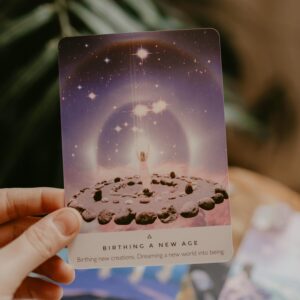 Tarot card held in hand, surrounded by crystals and plants, indoors.